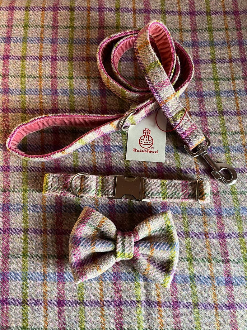 May include: A dog leash, collar, and bow tie set. The set features a plaid pattern in pink, green, yellow, and blue. The leash has a pink interior and a metal clasp. The collar has a metal buckle. The bow tie is pre-tied.
