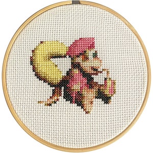 Może przedstawiać: Haft krzyżykowy przedstawiający postać z kreskówki w drewnianej tamborku. Postać ma żółty ogon, różową czapkę oraz różowo-brązowy strój. Tło to biała tkanina z wzorem kratki.