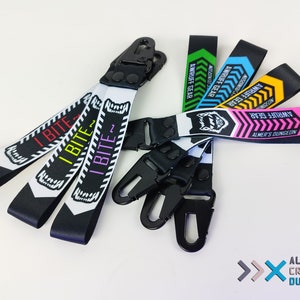 以下が含まれることがあります： 黒と白のデザインが施されたカラフルなキーホルダーのセット。キーホルダーには「I BITE」と「AWRUFF GEAR」の文字が書かれています。1つのキーホルダーはピンク、黒、白のデザインで、「ALMER'S DUNGEON」の文字が書かれています。