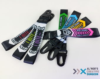 Può includere: Un set di portachiavi colorati con disegni in bianco e nero. I portachiavi presentano il testo "I BITE" e "AWRUFF GEAR". Un portachiavi ha un design rosa, nero e bianco con il testo "ALMER'S DUNGEON".
