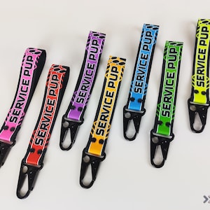 May include: Six colourful keychains with black metal clasps. Each keychain has the text "SERVICE PUP" printed on it in black and white. The keychains are in the colours pink, red, purple, yellow, blue, and green.
