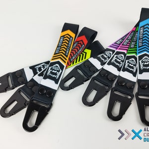 Könnte beinhalten: Ein Set mit sechs bunten Schlüsselanhängern mit einem schwarz-weißen Wolf-Design. Jeder Schlüsselanhänger hat einen schwarzen Metallclip und den Text "AWRUFF GEAR" und "ALMER'S DUNGEON".