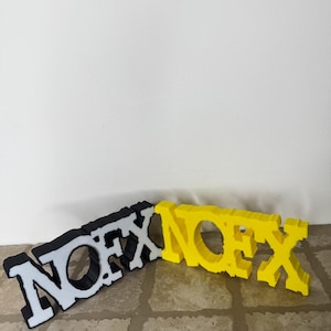 Könnte beinhalten: Zwei 3D-gedruckte NOFX-Logos, eines in Schwarz mit weißen Buchstaben und eines in Gelb. Die Logos liegen auf einer gefliesten Oberfläche.