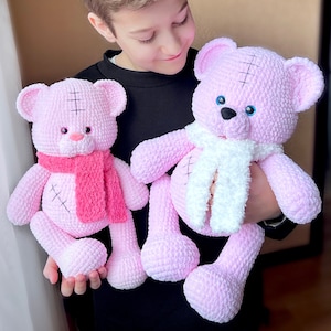 Op de afbeelding: Twee roze gehaakte teddyberen met witte en roze sjaals. De beren hebben blauwe ogen en gestikte neuzen. Een jong persoon houdt de beren vast.
