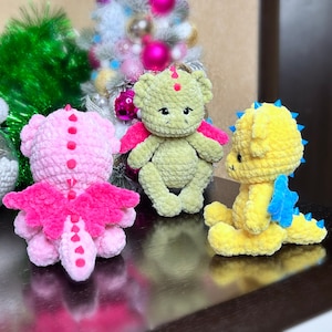 Peut inclure: Quatre jouets en forme de dragon en crochet de différentes couleurs. Un dragon rose avec des ailes roses, un dragon vert avec des ailes roses, un dragon jaune avec des ailes bleues et un bonnet de Père Noël rouge et blanc.