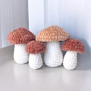 May include: A collection of four crochet mushrooms in varying sizes. The mushroom caps are in shades of brown and tan, while the stems are white. The textured yarn gives a soft, plush appearance. These decorative items are likely handmade.