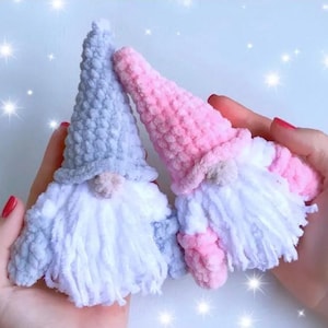 May include: Two crocheted gnomes, one gray and one pink, with white yarn beards. The gnomes are held in a person's hands.