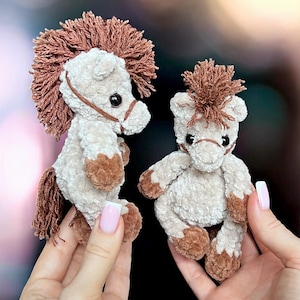 May include: Two small, plush, cream-colored crocheted horses with brown manes, tails, and hooves. Each horse features a brown yarn bridle and dark, round eyes. The horses are crafted from a soft, textured yarn material with a fuzzy appearance.