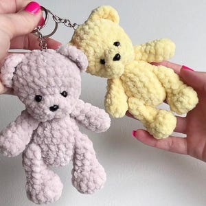 May include: Two handmade crochet teddy bear keychains. One is light purple, the other is yellow. Both have black button eyes and black stitched noses. The bears are soft and fluffy, with a keychain ring attached to the purple bear.
