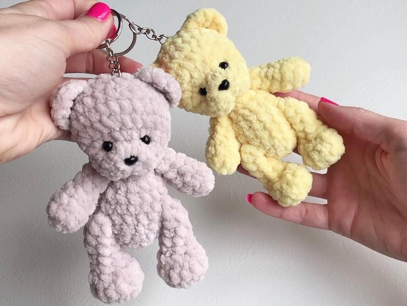 May include: Two handmade crochet teddy bear keychains. One is light purple, the other is yellow. Both have black button eyes and black stitched noses. The bears are soft and fluffy, with a keychain ring attached to the purple bear.