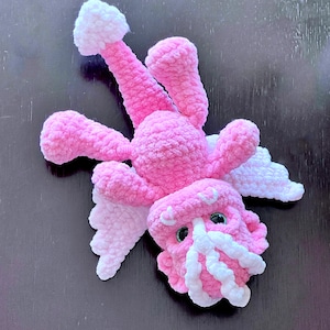 Może przedstawiać: Różowy szydełkowy smok z białymi skrzydłami i białą koroną. Smok ma duże zielone oczy i różowo-biały szydełkowy ogon.