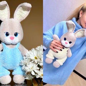 May include: A crocheted bunny plush toy with blue overalls. The bunny has a light brown body, white ears, and blue eyes. The bunny is sitting on a white surface with a bouquet of white flowers.