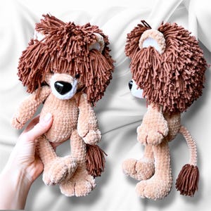 Puede incluir: Un león de peluche hecho de hilo suave de color marrón claro con una melena texturizada y desgreñada de color marrón oscuro. El león presenta un hocico blanco, una nariz negra en forma de corazón y una cola larga con un mechón de hilo.