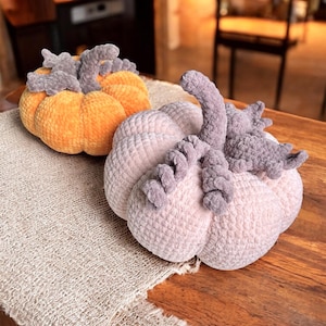 Op de afbeelding: Twee pluche pompoenen in oranje en roze tinten liggen op een houten tafel. De pompoenen hebben grijze stelen en zijn gemaakt van een zacht, getextureerd materiaal. Onder de pompoenen ligt een beige jute loper.