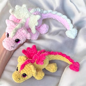 May include: Two crocheted dragon plushies. One is pink with white wings and a white tail tip. The other is yellow with pink wings and a pink tail tip.