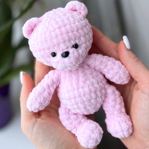 May include: A small pink crocheted teddy bear with black eyes. The bear is held in a person's hand.