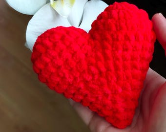 May include: A vibrant red, heart-shaped crochet item. The handmade piece is held in a hand, with a white orchid flower in the background. The heart has a textured, plush appearance, suggesting a soft, tactile quality. The image is well-lit, highlighting the color and texture.