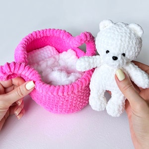 May include: A white crocheted teddy bear sitting in a pink crocheted basket. The basket is filled with white crocheted stuffing.