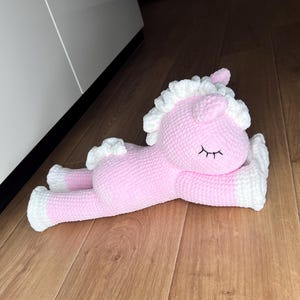 Puede incluir: Un juguete de peluche con forma de unicornio hecho de hilo suave rosa y blanco. El unicornio tiene un cuerpo rosa, pezuñas blancas, una crin blanca y un cuerno pequeño. Está en posición tumbada con ojos cerrados bordados en hilo negro.