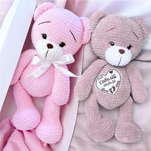 Puede incluir: Dos osos de peluche de ganchillo hechos a mano. Uno es rosa claro con un lazo blanco, el otro es malva con un parche en forma de corazón. El parche tiene texto y un diseño de huella. Ambos osos tienen narices marrones y ojos negros.
