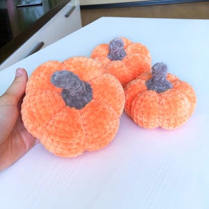 May include: Three plush orange pumpkins with gray stems are arranged on a white surface. The pumpkins vary in size, with the largest one in the foreground. The soft, textured material gives them a cozy, handmade appearance, perfect for fall decor.