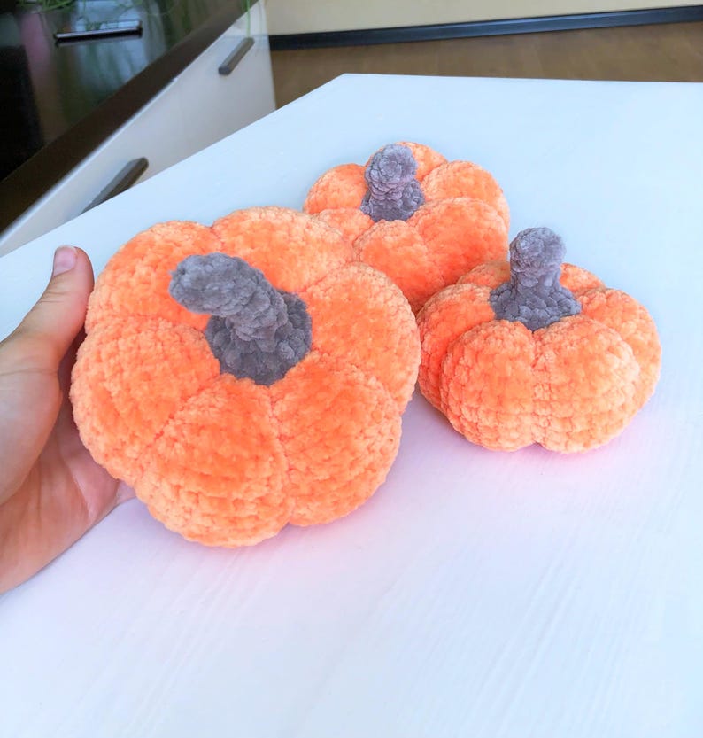 May include: Three plush orange pumpkins with gray stems are arranged on a white surface. The pumpkins vary in size, with the largest one in the foreground. The soft, textured material gives them a cozy, handmade appearance, perfect for fall decor.