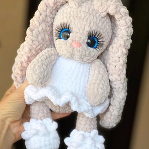 May include: A plush crochet bunny toy made of soft beige yarn with long ears. The bunny features large blue eyes with prominent black eyelashes, a small pink nose, and is wearing a white crocheted dress with a ruffled hem and white booties.