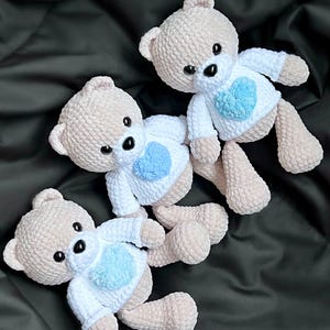 May include: Three crocheted teddy bears with white shirts and blue hearts. The bears are sitting on a black fabric background.