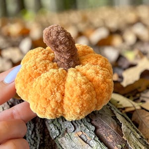 May include: A small, round, orange crocheted pumpkin with a textured, fuzzy surface and a thick, brown, upright stem. The pumpkin features distinct, segmented ridges created by the crochet stitching, giving it a soft, plush, and rounded appearance.