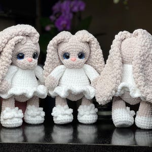 Könnte beinhalten: Drei handgefertigte Häkel-Plüschhasen mit langen, hängenden Ohren. Jeder Hase hat einen hellbeigen Körper, ein weißes Kleid mit gerüschtem Rock und weiße Rüschenschuhe. Sie haben große, runde blaue Augen mit langen Wimpern und eine kleine rosa Nase.