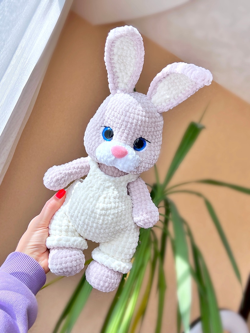 May include: A crocheted plush bunny toy with white overalls, light purple fur, and blue eyes. The bunny is being held by a hand.