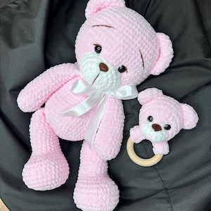 Pode incluir: Um urso de pelúcia rosa de crochê com uma fita branca ao redor do pescoço, ao lado de um urso rosa menor com um anel de madeira. Os ursos têm narizes marrons e olhos pretos. O urso maior tem cerca de 30 cm de altura.