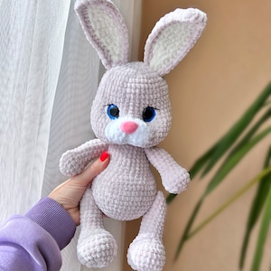 May include: A crocheted plush bunny toy with a light purple body, white ears, and blue eyes. The bunny has a pink nose and a white patch on its chest.
