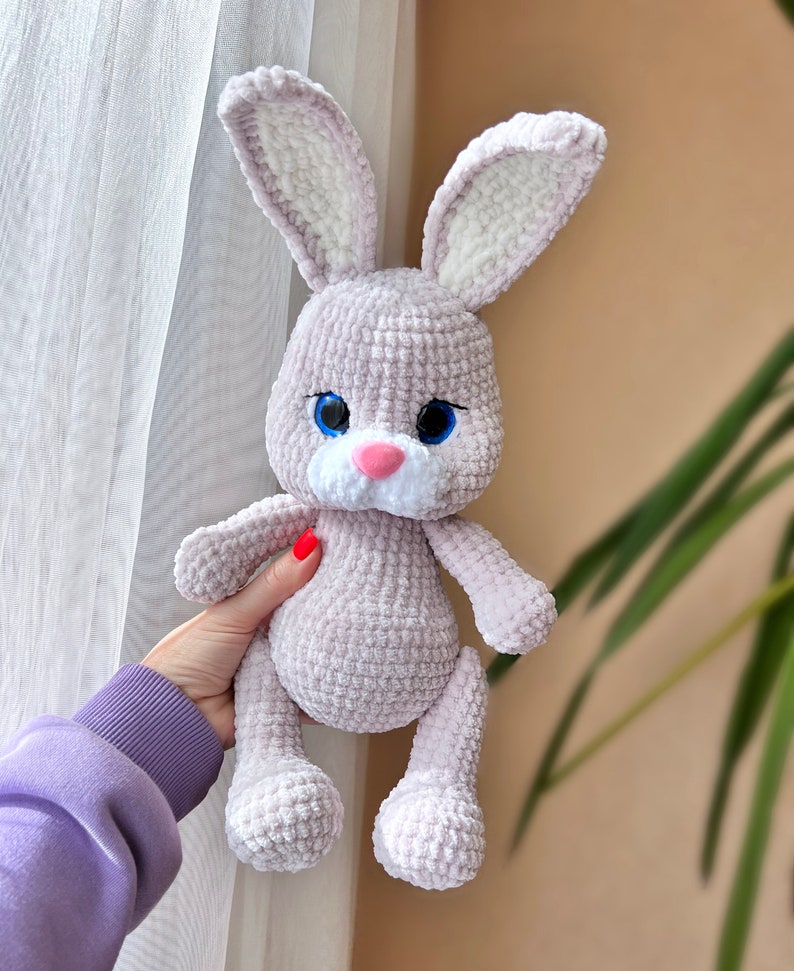 May include: A crocheted plush bunny toy with a light purple body, white ears, and blue eyes. The bunny has a pink nose and a white patch on its chest.