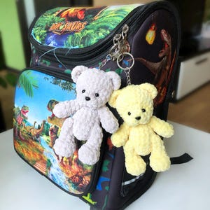 May include: A black backpack with a colorful dinosaur theme, featuring the word "DINOSAURS". Two small, handmade teddy bear keychains in light gray and yellow are attached to the zipper.