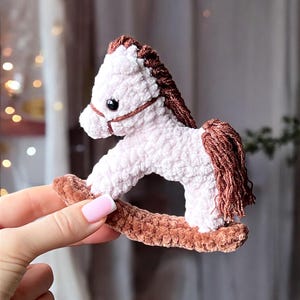Puede incluir: Un caballo balancín de peluche hecho a mano, rosa y marrón. El caballo presenta una melena y cola detalladas, con una brida marrón y una base curva. El juguete está diseñado para niños.