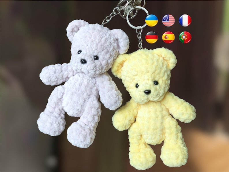 May include: Two plush teddy bear keychains. One is a soft lavender color, and the other is a pale yellow. Both have black button eyes and noses. The keychains are attached to a silver metal ring. Flags of different countries are in the background.