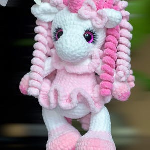 May include: A pink and white crocheted unicorn toy with pink yarn hair and a pink dress. The unicorn has sparkly pink eyes.
