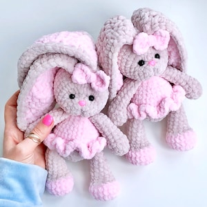 May include: Two plush, handmade bunny dolls. The bunnies are gray with pink accents, including the inner ears, bows, and dresses. They have black eyes and pink noses. The dolls are soft and fluffy, perfect for a child's toy collection.