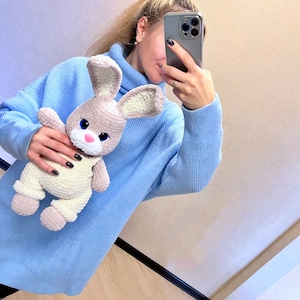 May include: A woman wearing a light blue turtleneck sweater is holding a crocheted light brown and white bunny rabbit toy.