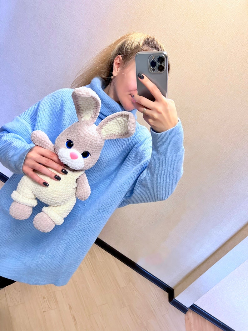 May include: A woman wearing a light blue turtleneck sweater is holding a crocheted light brown and white bunny rabbit toy.