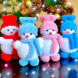 Op de afbeelding: Vier handgemaakte gehaakte sneeuwmannen in rood, blauw en roze. Elke sneeuwman heeft een bijpassende hoed, sjaal en voeten. Ze hebben oranje wortelneuzen en zwarte knoopogen. De sneeuwmannen zijn wit met kleurrijke accenten.