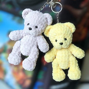 May include: Two handmade crochet teddy bear keychains. One is light lavender, the other is yellow. Both have black button eyes and are attached to silver keyrings. The bears are made of a soft, textured yarn.