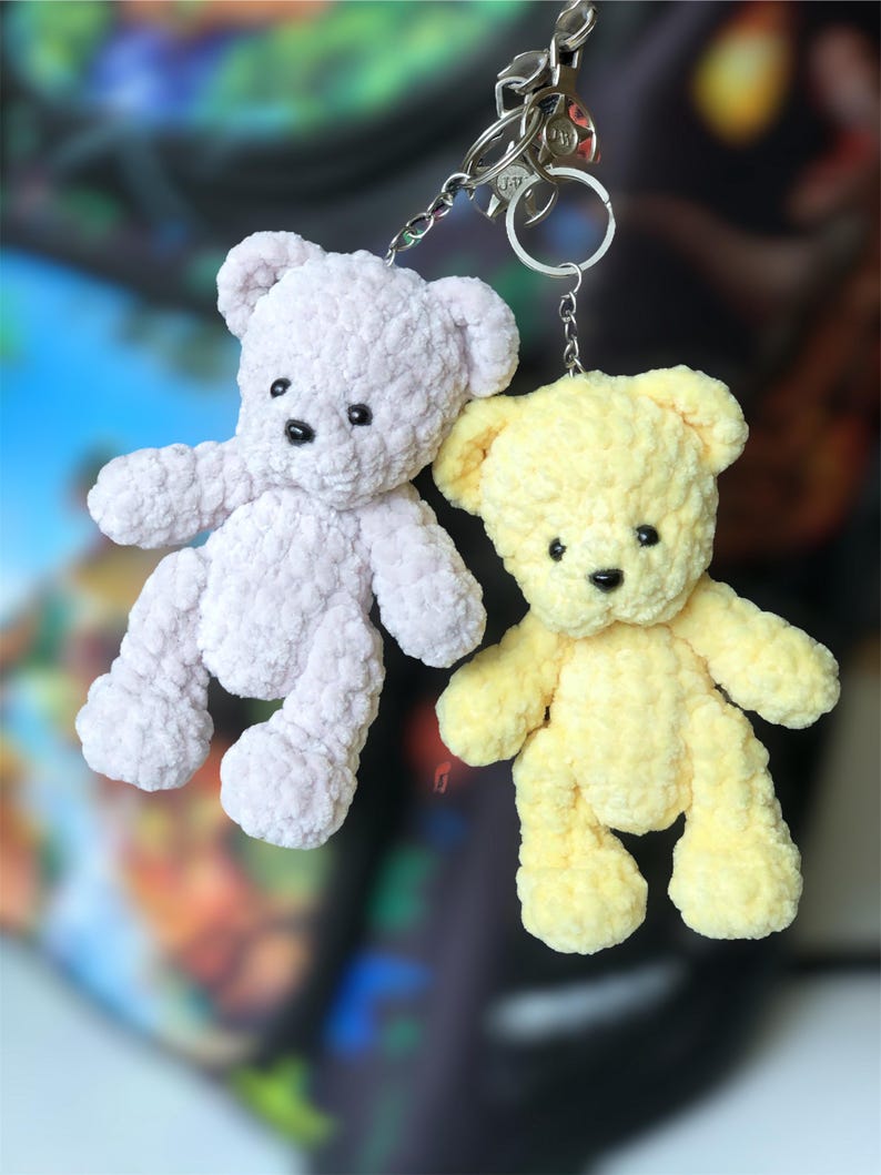 May include: Two handmade crochet teddy bear keychains. One is light purple, the other is yellow. Both have black eyes and are attached to silver keyrings. The bears are made of a soft, textured yarn.