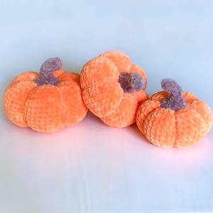 May include: Three plush orange pumpkins with gray stems are arranged on a white surface. The pumpkins have a soft, textured appearance, suggesting a handmade or crafted item. The pumpkins are likely decorative items for the fall season.