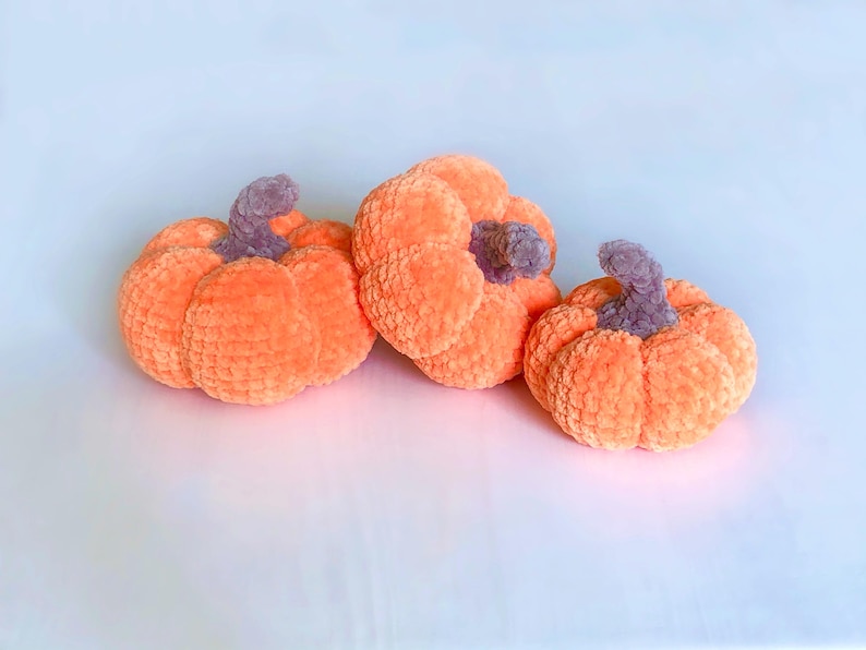 May include: Three plush orange pumpkins with gray stems are arranged on a white surface. The pumpkins have a soft, textured appearance, suggesting a handmade or crafted item. The pumpkins are likely decorative items for the fall season.