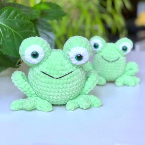 May include: Two handmade crochet frogs in a light green color. The frogs have large, round eyes with white and green details, and a smiling mouth. The frogs are sitting on a white surface with green foliage in the background.