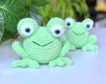 May include: Two handmade crochet frogs in a light green color. The frogs have large, round eyes with white and green details, and a smiling mouth. The frogs are sitting on a white surface with green foliage in the background.
