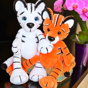 May include: Two crocheted tiger plushies, one white with black stripes and blue eyes, the other orange with black stripes, sitting together on a woven basket.