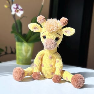 May include: A handmade yellow crochet giraffe with brown spots, ears, and hooves. The plush toy has a soft, textured appearance and is sitting on a white surface. The giraffe has a sweet expression.